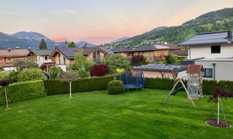 A beautiful garden with green grass and play equipment. In the background, houses and mountains are visible at sunset.