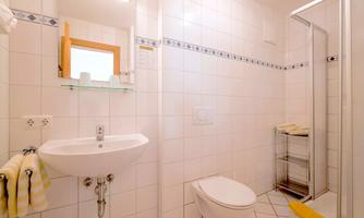 A modern bathroom with white tiles and a shower cabin. There is a sink, a toilet, and a shelf for towels.