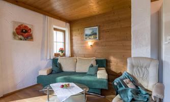 A cozy living room with a green sofa and a beige armchair. The walls are made of wood, and the light is warm and inviting.
