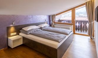 A modern bedroom with two single beds and large windows. The room has a warm wood floor and a cozy atmosphere.