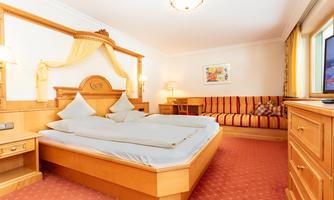 A cozy hotel room with a large bed and stylish wooden furniture. The walls are light, and there is a sofa as well as a window with a view outside.