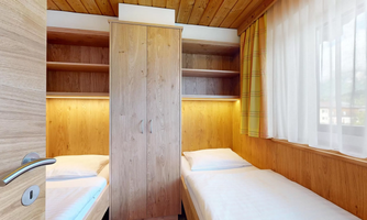 A cozy room with two single beds and a wooden wardrobe. The walls are paneled with light wood and there are many windows for natural light.