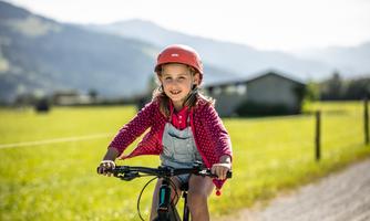 A girl rides a bicycle on a green field. She is wearing a helmet and is smiling kindly at the camera.
