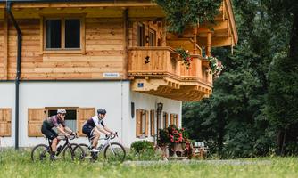 Two cyclists ride past a wooden house. Around the house, green meadows and flowers can be seen.