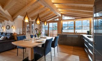 A modern living room with wooden ceilings and large windows. The dining area is stylishly furnished with a table and blue chairs.