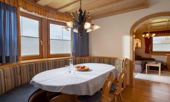 A cozy dining area with a round table and wooden chairs. Large windows and simple furniture create an inviting atmosphere.