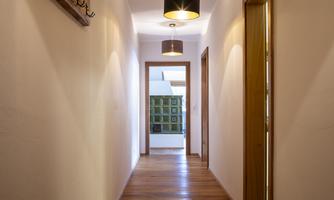 A bright hallway with wooden flooring and modern ceiling lights. At the end of the hallway, doors and windows are visible.