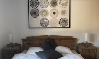 A cozy bedroom with a large bed and pillows in neutral colors. A modern artwork with circular patterns hangs on the wall.