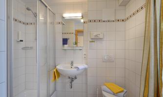 A modern bathroom with a shower, a sink, and a toilet. The walls are tiled, and there is a large mirror as well as yellow towels.