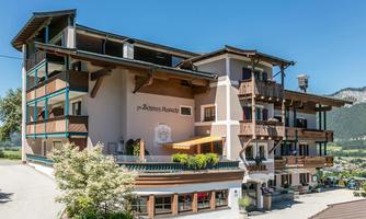 A charming hotel building with several balconies and an inviting terrace. The surroundings are green and offer a beautiful view of the mountains.