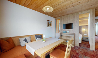 A cozy living space with a wooden table, chairs, and a seating area. A television and a picture hang on the wall, while a flower adorns the table.