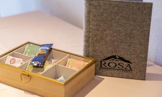 A decorative wooden box with various tea bags and a small snack. Next to it is a gray menu book with the print "ROSA".