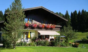 A charming country house with blooming balcony boxes and a green garden. In the background, tall trees and a blue sky can be seen.