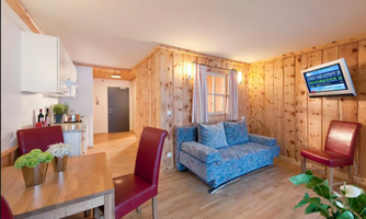 A cozy living space with wooden walls, a blue couch, and a dining table. The windows let in plenty of daylight, and the television is mounted on the wall.
