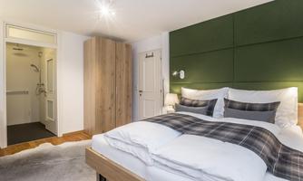 A cozy bedroom with a large bed and a green wall. The room is modernly furnished and has access to a bathroom.