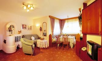 A cozy living room with a green sofa and a dining table. The room design is friendly and inviting, with carpet flooring and warm colors.