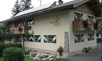 A cozy house with a balcony, surrounded by greenery and flowers. The facade is bright with typical wooden details.