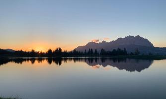 A tranquil lake at sunset with mountains in the background. The sky is colorful and reflected in the water.