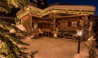 A charming alpine hut with festive lighting and a snow-covered ground. Surrounded by pine trees, it exudes a cozy winter atmosphere.