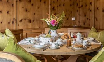 A beautifully set table in a wooden room. It is decorated with a flower vase, dishes, and fresh rolls.