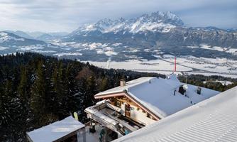 A snow-covered alpine hut overlooking the mountains and a deep valley. The landscape is characterized by white snow and green forest areas.