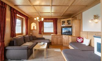 A cozy living room with wooden walls and large windows. There is a comfortable couch, a TV cabinet, and decorative elements.