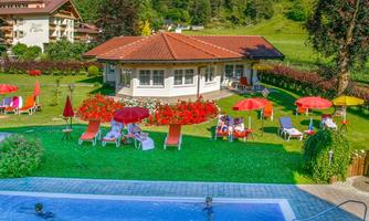 A beautiful pool surrounded by a green meadow and colorful flowers. In the background, mountainous landscapes and cozy buildings can be seen.