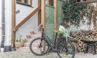 A stylish bicycle stands in front of a beautiful house with wooden steps. Plants and firewood add a cozy atmosphere to the scene.