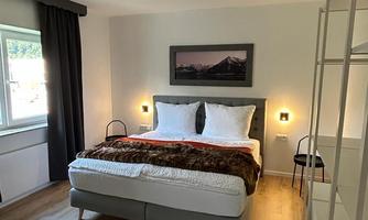 A modern bedroom with a double bed and a soft bedspread. The walls are light, and a window lets in daylight.