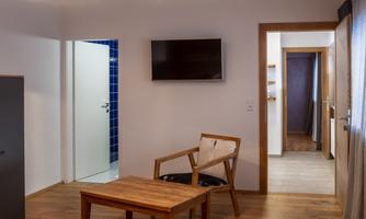 A cozy room with wooden floors, a table, and a chair. A television is mounted on the wall, and there are two doors leading to additional rooms.