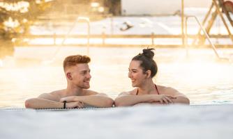 A couple relaxes in the outdoor pool in a wintry setting. In the background, snow-covered mountains and a wooden building can be seen.