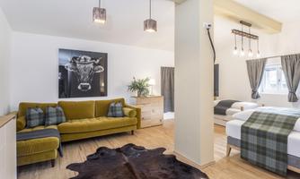 A modern room with a green sofa, two single beds, and a stylish cow portrait on the wall. The floor is covered with a decorative rug and there are large windows with curtains.