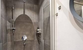 A modern shower with gray tiles and glass sliding doors. In the background, a round mirror is visible.