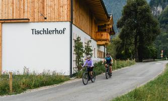 Two cyclists ride along a quiet path by a wooden house. In the background, mountains and trees are visible.