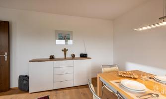 A bright dining room with a wooden table and modern chairs. There is an elegant sideboard with decoration on the wall.