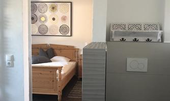 A modern bathroom with a toilet area and an adjoining bedroom. The decor is simple and stylish, with wood and neutral tones.