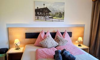 A cozy bedroom with a large bed and checkered pillows. On the wall hangs a picture of an idyllic mountain landscape.
