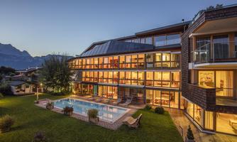 A modern hotel building with large windows and a clear pool in the foreground. In the background, mountains and a well-kept garden can be seen.