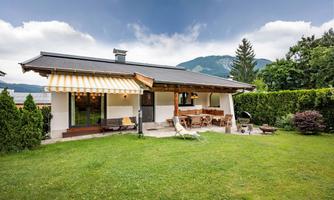 A charming house with a well-maintained garden and a terrace. In the background, green mountains and a cloudy sky can be seen.