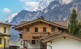 A rustic wooden house in the mountains with snow-covered peaks in the background. The surroundings are green and inviting.