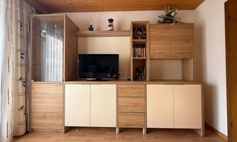 A modern living room with a wooden shelf and a television. The walls are light and there are large windows with curtains.