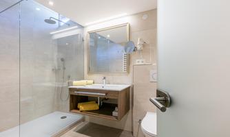A modern bathroom with a walk-in shower and a wooden vanity. There are bright tiles and ample mirror lighting.