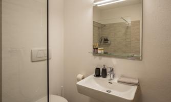 A modern bathroom with a shower, sink, and mirror. The walls are bright, and the furnishings are simple and functional.