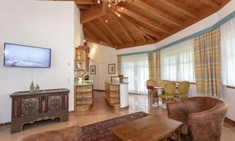 A cozy living room with a wooden beam ceiling and large windows. The furnishings include a television, a dining table, and comfortable armchairs.
