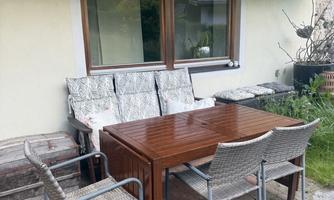 A cozy seating area outdoors with a wooden table and rattan chairs. In the background, a large window and some greenery can be seen.