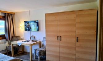 A modern room with a double bed, a large wardrobe, and a table. A television is mounted on the wall, and there are two cozy chairs.