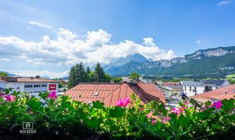 A picturesque view of mountains and houses under a blue sky. Colorful flowers in a flowerpot in the foreground add a vibrant accent to the image.