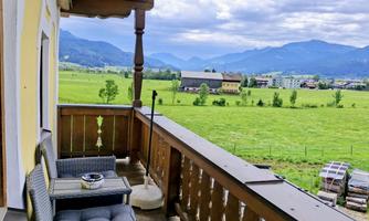 A cozy balcony with two chairs and a table. In the background, a green meadow stretches out and the mountains under a cloudy sky.