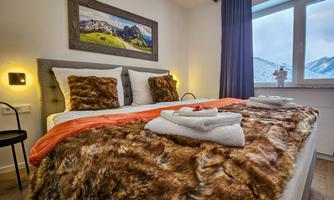 A cozy bedroom with a large bed and a soft, fur-like throw. An alpine landscape is visible through the window.