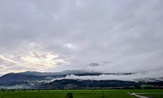A picturesque landscape with mountains in the background and low-hanging clouds. In the foreground, a green meadow stretches along a winding road.
